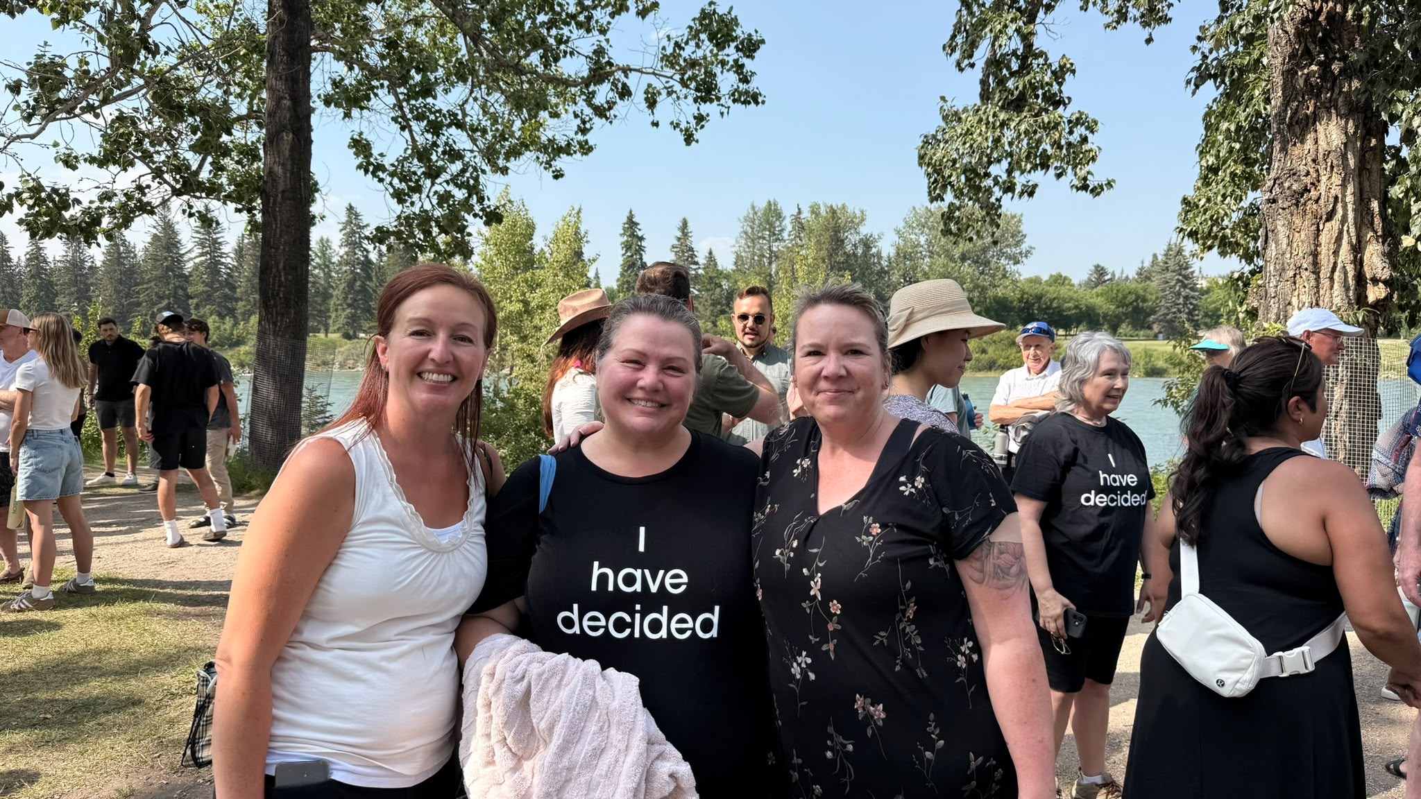 Three women smiling together on a riverbank in Bowness, Calgary; the woman in the center wears a black "I have decided" T-shirt and holds a towel, with a crowd of people and the Bow River behind them.