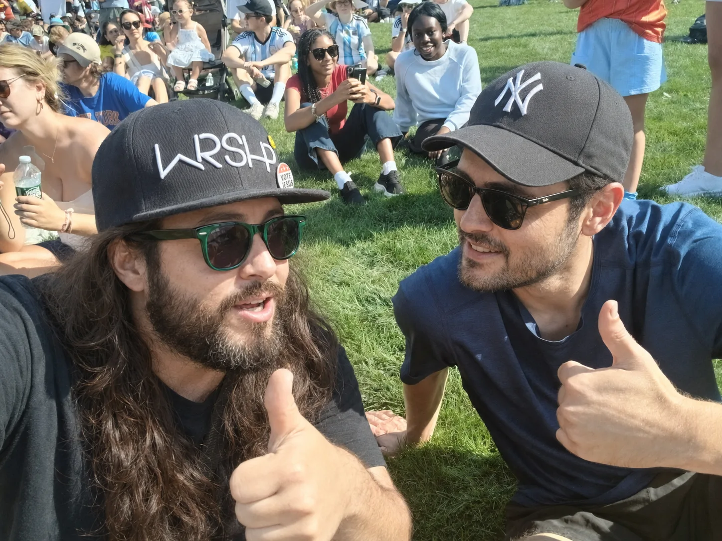 Two men sit on the grass at a crowded outdoor gathering, talking, one giving a thumbs up.