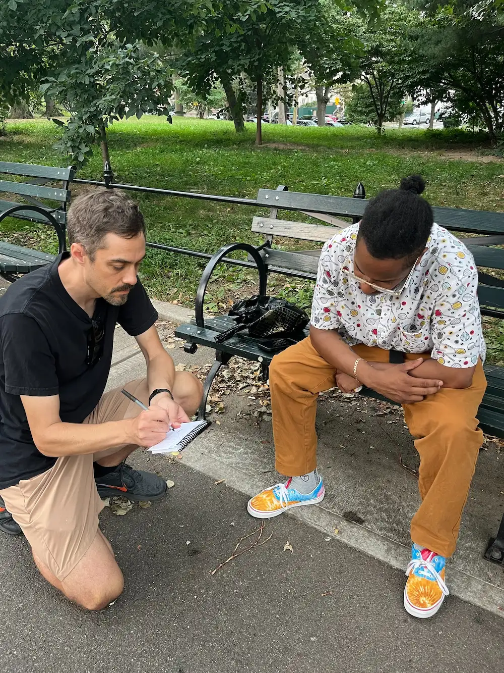 A man kneels on a park path sketching in a small notebook while another man leans in from a green bench to watch.
