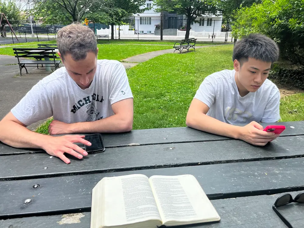 Two young men sit at a park picnic table with an open Bible in front of them, each looking down at a phone.