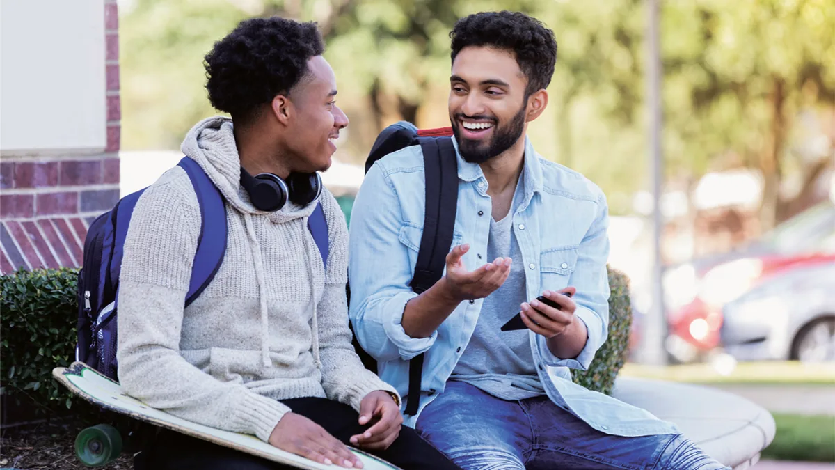Two young men sit talking and smiling on a bench, one holding a phone